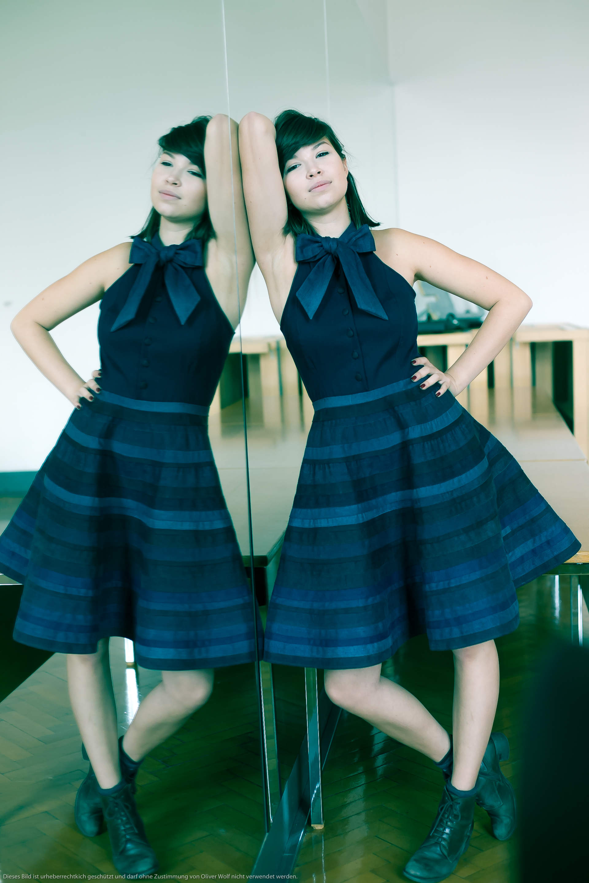 Eine Frau in einem marineblauen Kleid mit einer großen Schleife steht mit erhobenen Armen vor einem Spiegel. Sie trägt Stiefeletten und blickt mit leicht geneigtem Kopf in die Kamera. Im Hintergrund ist ein Raum mit Holzmöbeln zu sehen.