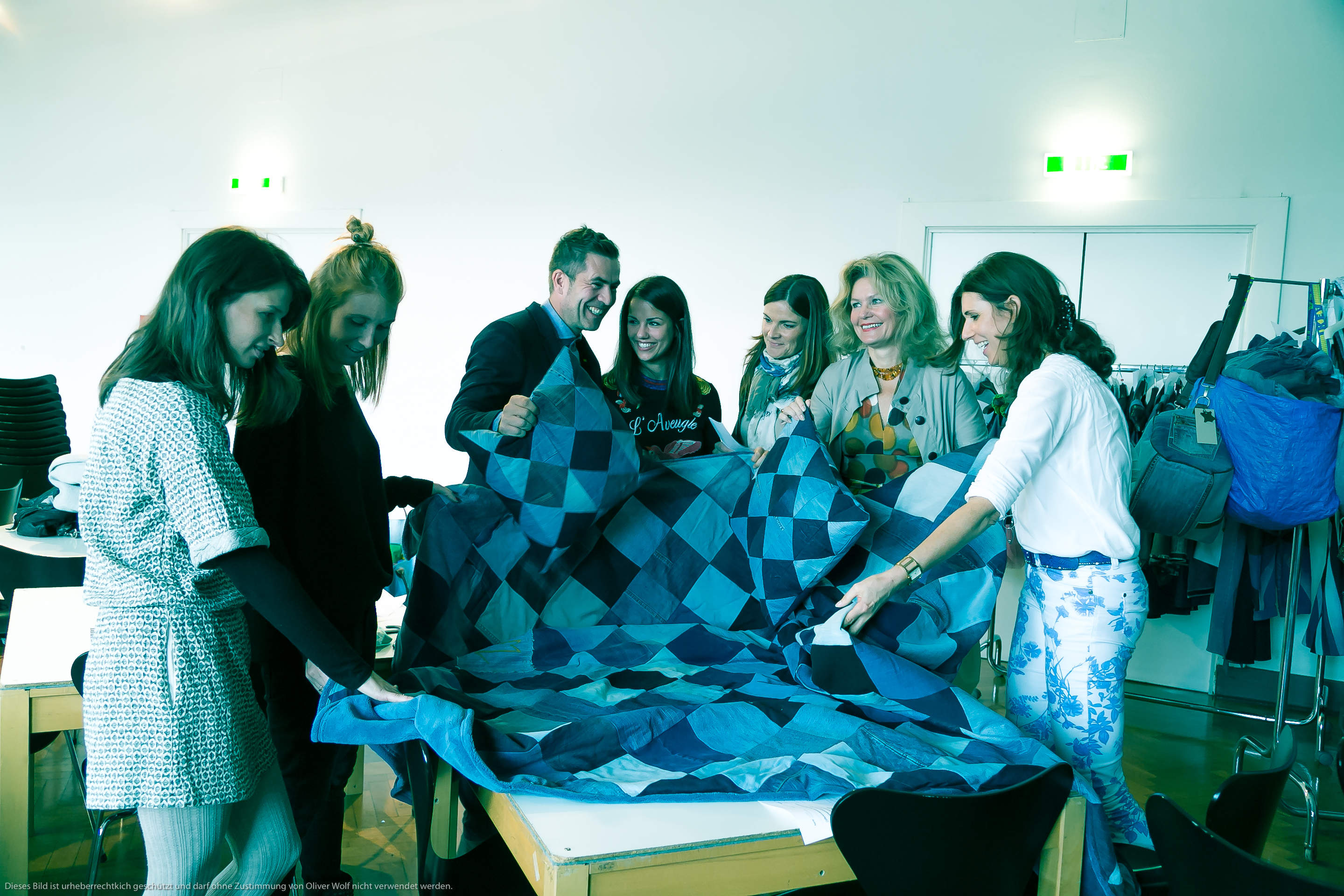 Eine Gruppe von Menschen steht lächelnd um einen Tisch herum und hält eine große, blaue Steppdecke mit geometrischem Muster in den Händen. Sie scheinen sich in einem gut beleuchteten Raum zu befinden, im Hintergrund stehen Kleiderständer.