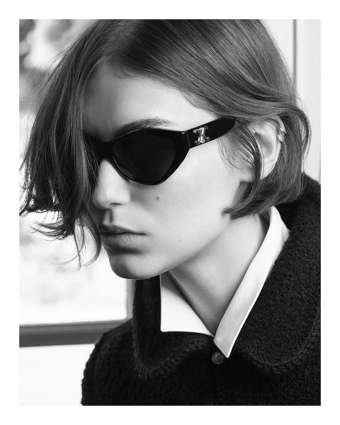 Photo en noir et blanc d'une personne aux cheveux courts portant des lunettes de soleil foncées et une chemise à col sous une veste texturée, regardant sur le côté dans un environnement faiblement éclairé.