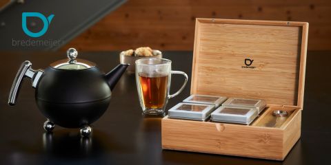 Sur une table sombre se trouvent une théière noire, un gobelet en verre rempli de thé et une boîte en bois contenant quatre boîtes de thé. Le logo "Bredemeijer" est visible sur la théière et la boîte.