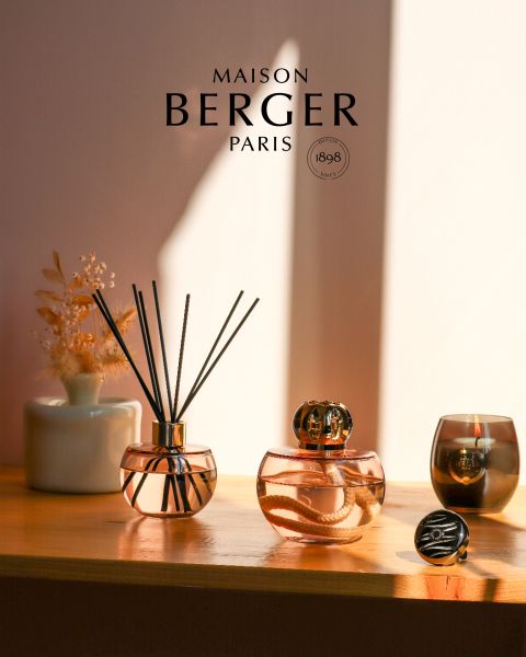 Sur une étagère ensoleillée, deux diffuseurs de Maison Berger Paris au design élégant en verre. Un diffuseur est équipé de tiges de rotin, l'autre d'un bouchon décoratif. À côté, un petit vase avec des fleurs séchées et une bougie créent une atmosphère chaleureuse et accueillante.