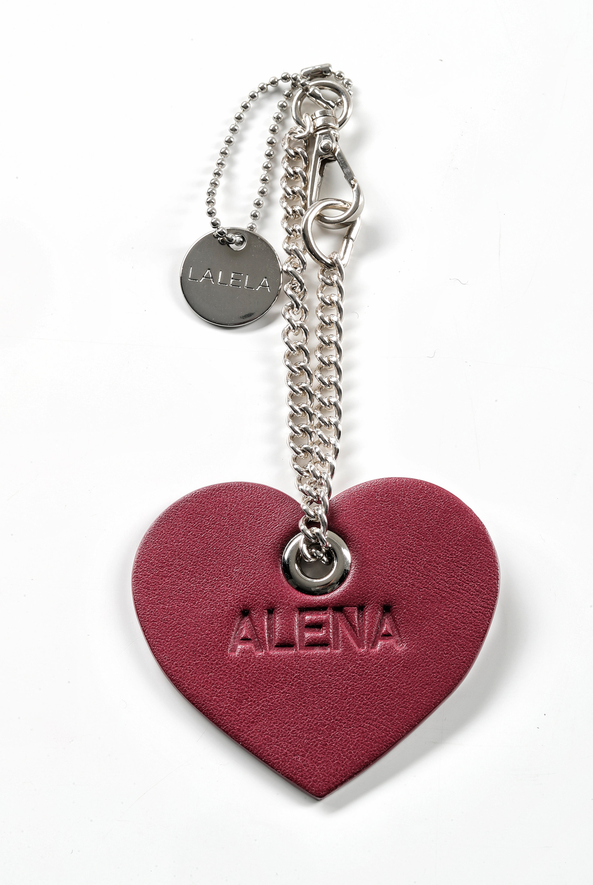 Ein herzförmiger Schlüsselanhänger aus burgunderfarbenem Leder mit dem eingeprägten Namen „ALENA“. An der Kette ist eine silberne Kugelkette mit einem Metallschild mit der Aufschrift „LAFELA“ und einem Karabinerverschluss befestigt.