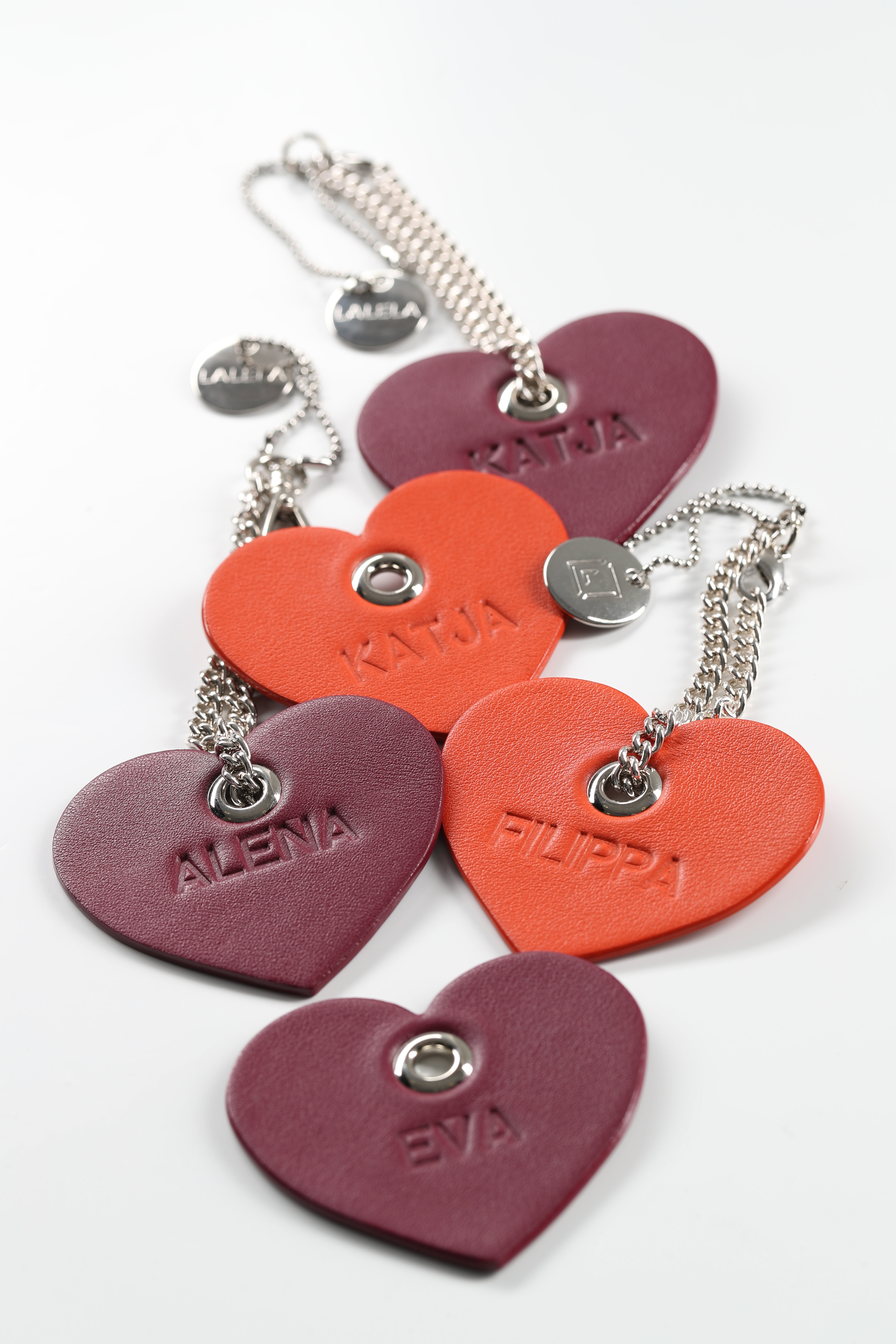 Herzförmige Schlüsselanhänger aus Leder in Rot-, Lila- und Orangetönen, auf denen jeweils ein anderer Name eingeprägt ist. Verbunden sind sie durch silberne Ketten und kleine runde Metallanhänger auf weißem Hintergrund.