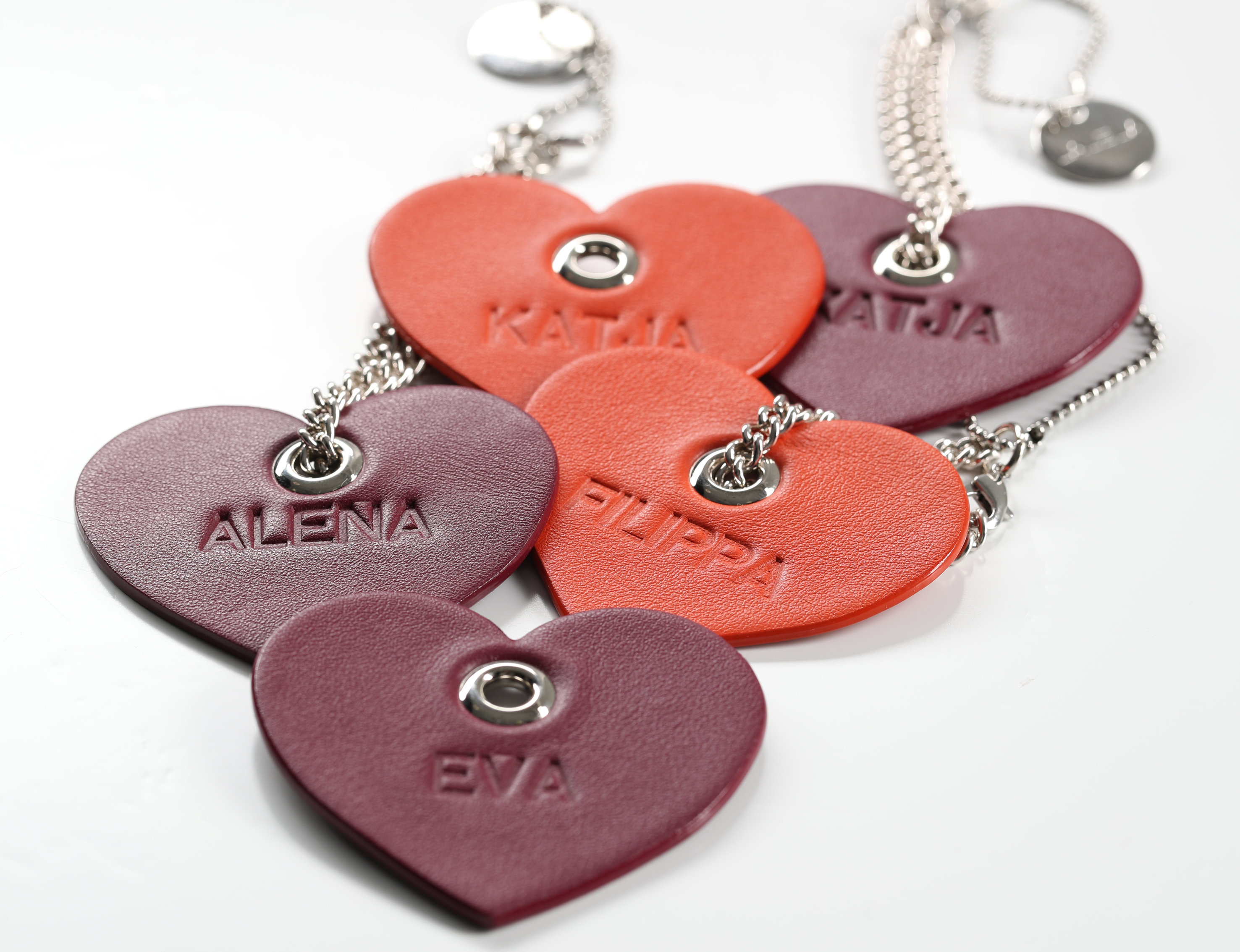 Herzförmige Schlüsselanhänger mit geprägten Namen: Katia und Katja in Orange, Alena, Filippa und Eva in Rot. Sie sind an silbernen Ketten befestigt und auf weißem Hintergrund angeordnet.