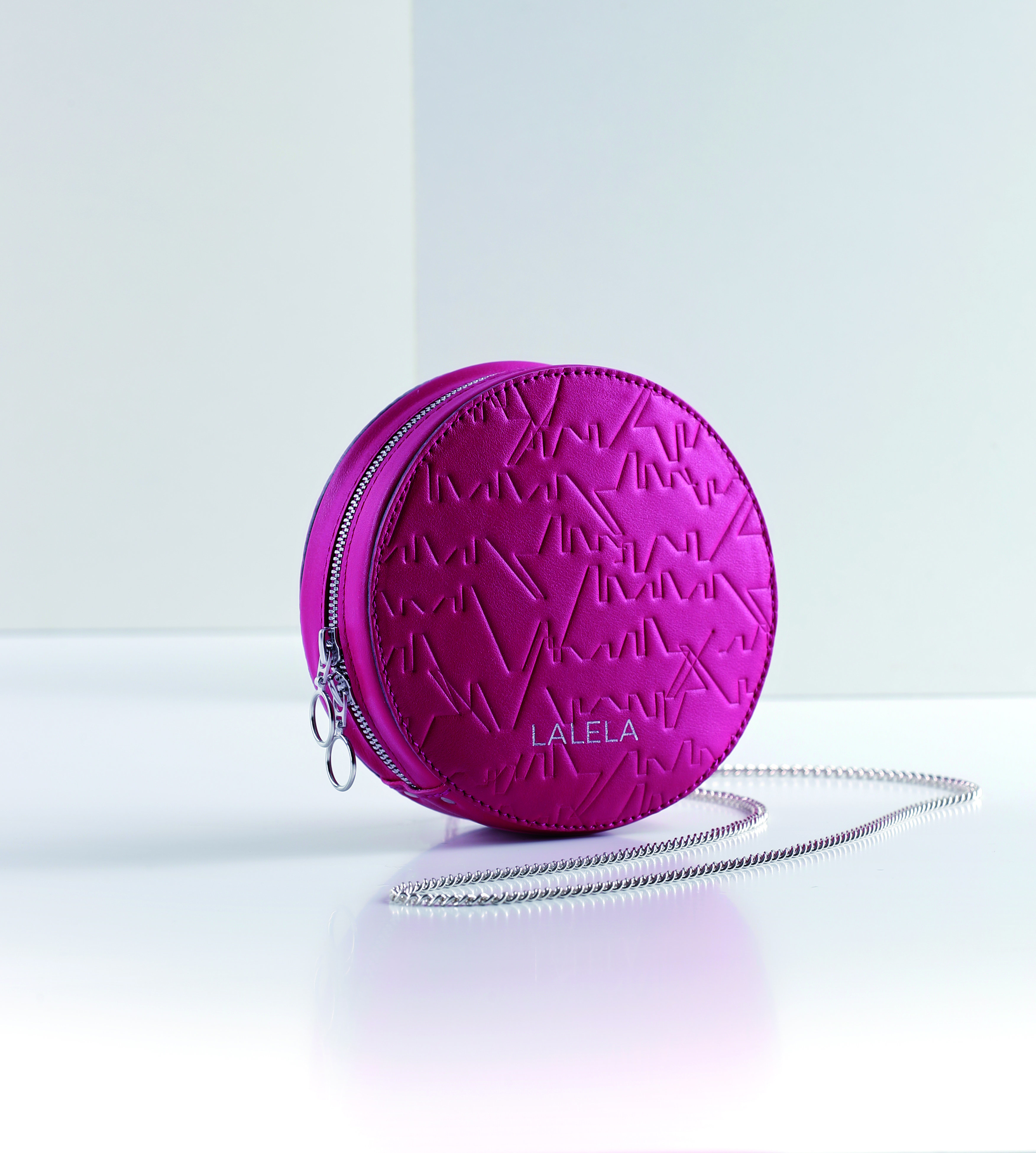 Eine runde, leuchtend violette Clutch mit strukturiertem Design und einer silbernen Kette auf einer glatten weißen Oberfläche. Der Markenname „LALELA“ ist auf der Vorderseite aufgedruckt. Die Tasche hat ein modernes, stilvolles Aussehen.