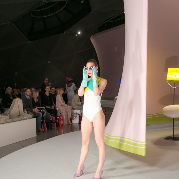 Un mannequin en maillot de bain blanc et gants bleus pose sur un podium futuriste. En arrière-plan, le public regarde. Le décor est composé de grandes structures géométriques et d'une lampe moderne.