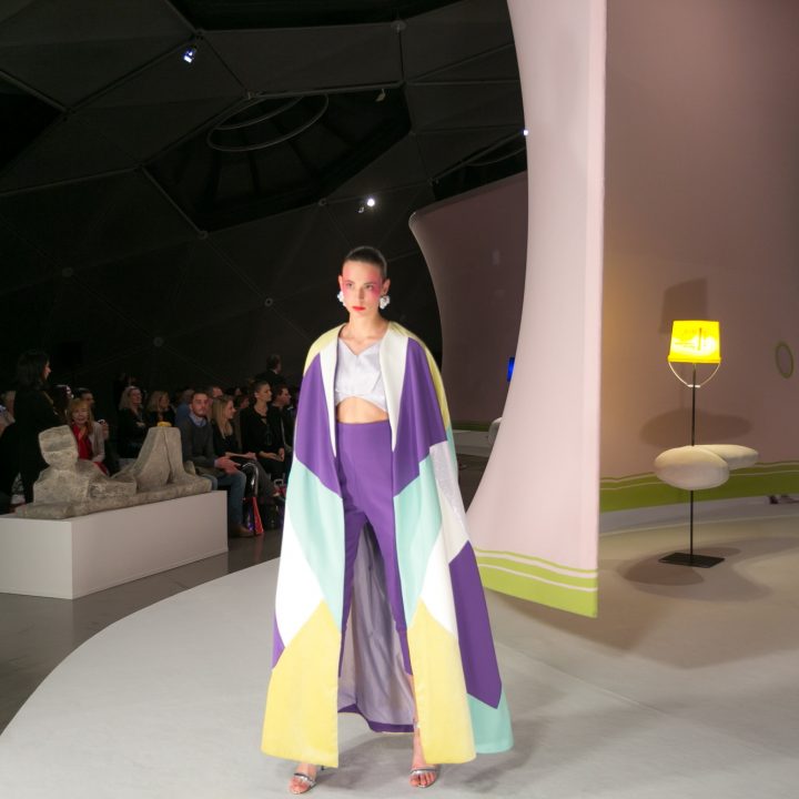 Un mannequin défile sur le podium dans une tenue colorée et fluide, ornée d'un motif géométrique violet, jaune et bleu. En arrière-plan, on aperçoit une installation artistique moderne aux formes sinueuses. Le public regarde dans la pièce faiblement éclairée.