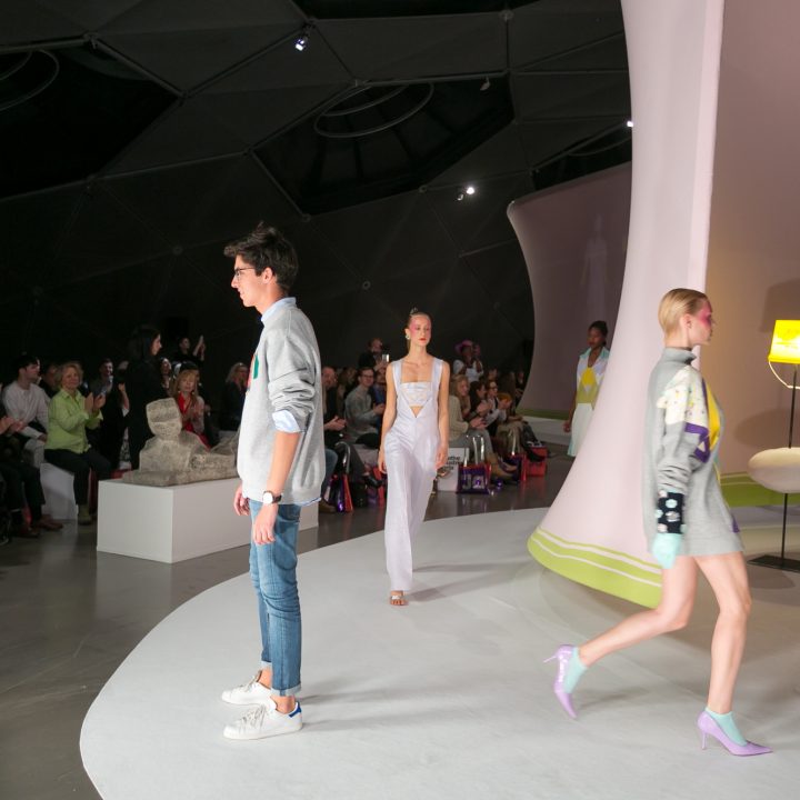 Des mannequins défilent sur le podium dans des tenues élégantes, dont un homme en pull décontracté et jean, et des femmes en robes élégantes. Le public regarde attentivement depuis le côté dans une ambiance moderne avec un éclairage doux.