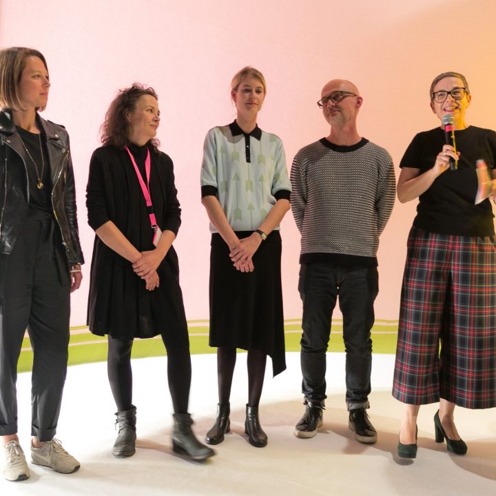 Cinq personnes sont sur scène sous une lumière vive. Quatre sourient et une parle dans un microphone. Ils sont vêtus de façon décontractée, avec une toile rose clair en arrière-plan et quelques spectateurs assis.