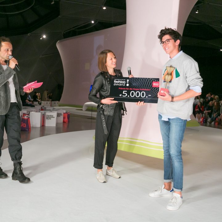 Un homme sur scène remet un prix de la mode à un récipiendaire souriant, qui tient une grande carte de prix avec le numéro « 5 000 ». Une autre personne se tient à proximité avec un microphone. La scène est lumineuse, avec un public assis et un décor moderne en arrière-plan.