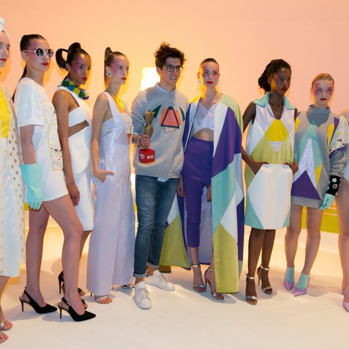 Un groupe de mannequins présente des créations de mode géométriques et colorées à côté d'une personne vêtue de manière décontractée. Les tenues se distinguent par des formes audacieuses et des couleurs vives. Les mannequins et la personne se tiennent devant un fond rose.