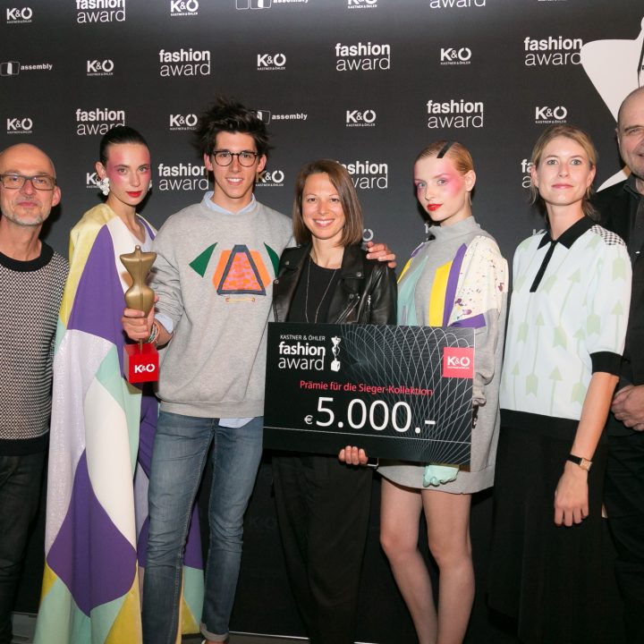 Un groupe de sept personnes sourient devant un fond noir avec des logos "Fashion Award". Une personne tient un trophée, une autre un grand chèque de 5 000 euros. Ils sont tous vêtus de vêtements élégants et colorés.