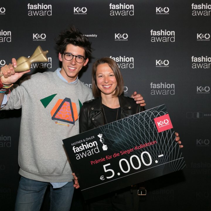 Deux personnes souriantes posent lors d'un événement de remise de prix de la mode. L'une tient un trophée, l'autre un grand chèque de 5 000 euros. Les deux se tiennent devant un fond avec les logos "Fashion Award" et "K&O".
