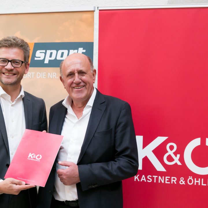 Deux hommes en costume sont debout, souriants. L'un d'eux tient un classeur rouge avec l'inscription "K&O Kastner & Öhler". Derrière eux, des bannières affichent les logos de Sky Sport et Kastner & Öhler.