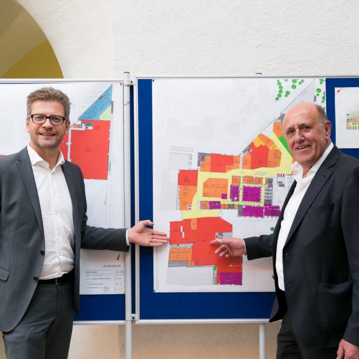 Deux hommes en costume se tiennent devant un tableau noir sur lequel sont accrochés des plans d'architecture colorés. Ils sourient tous les deux et gesticulent en direction des plans, ce qui suggère une discussion ou une présentation sur les projets.