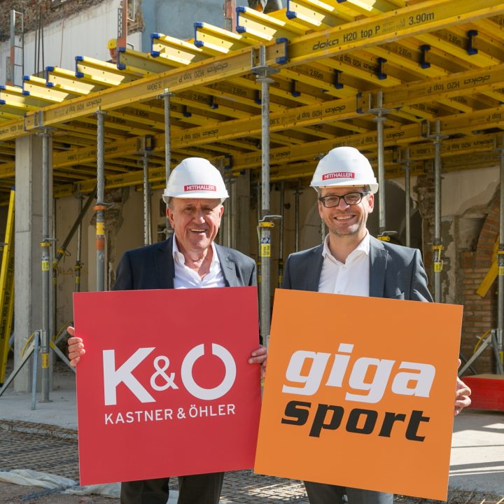 Deux hommes en costume et casque de chantier sourient et tiennent des panneaux pour Kastner & Öhler et Gigasport sur un chantier. Des échafaudages et des matériaux de construction sont visibles en arrière-plan.