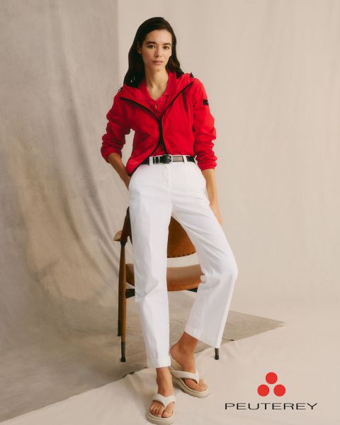 Une femme en veste rouge et pantalon blanc est appuyée contre une chaise. Elle a de longs cheveux foncés et porte des sandales blanches. La toile de fond est minimaliste avec un arrière-plan neutre. Le logo « Peuterey » se trouve dans le coin inférieur droit.