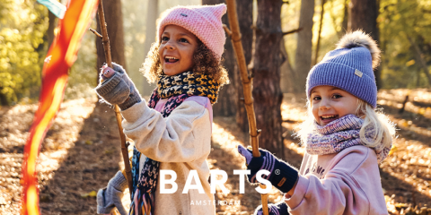 Deux enfants courent dans une forêt ensoleillée. Ils portent des bonnets d'hiver, des écharpes et des gants. Ils sourient et tiennent des bâtons avec des rubans colorés à la main. En bas de l'image, on peut lire le texte « BARTS AMSTERDAM ».