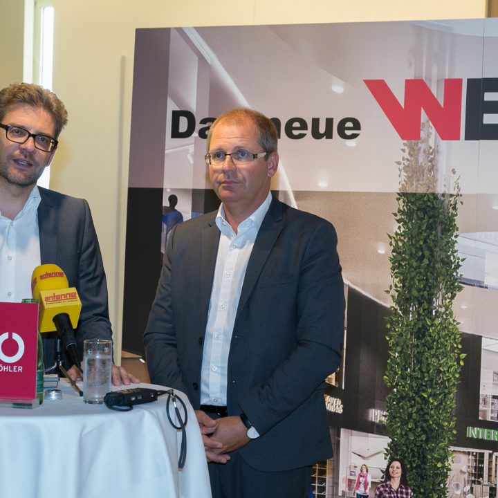 Deux hommes en costume se tiennent à une petite table ronde avec des microphones et s'expriment lors d'un événement. En arrière-plan, on peut voir « Le nouveau WEZ » avec des images de l'intérieur d'un centre commercial. Le décor semble formel, peut-être une conférence de presse ou une présentation.