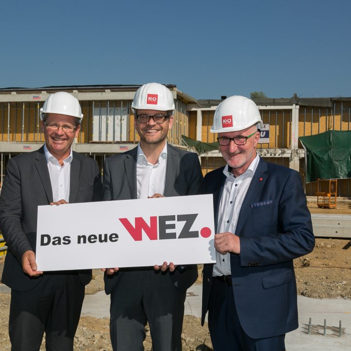 Trois hommes en costume et casque de chantier se tiennent souriants sur un chantier de construction et tiennent une pancarte indiquant « Le nouveau WEZ ». Derrière eux, on aperçoit un bâtiment à moitié terminé sous un ciel bleu éclatant.