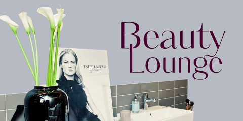 Une vitrine avec un vase noir contenant des fleurs blanches, une affiche d'une femme et les mots « Beauty Lounge » dans un texte élégant. La toile de fond comprend un mur carrelé avec des produits de beauté et des pinceaux de maquillage sur une surface à proximité.