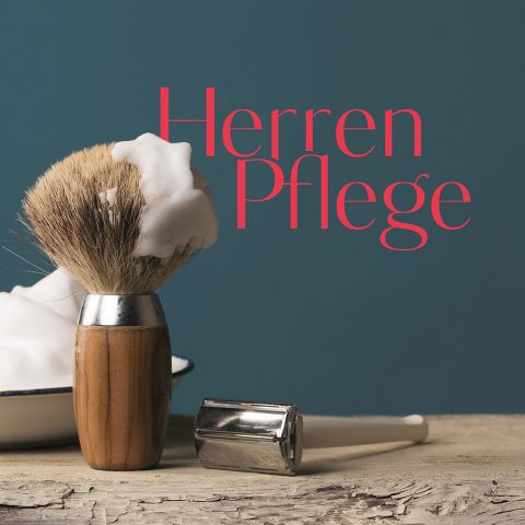 Un blaireau de rasage en bois avec de la mousse, un rasoir en métal et un bol sont posés sur une surface en bois devant un fond bleu. Au-dessus des articles, il est écrit en rouge « Soins pour hommes ».