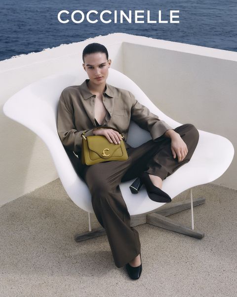 Une femme en tenue marron est assise sur une chaise blanche au bord de la mer et tient un sac à main Coccinelle jaune. Le logo Coccinelle se trouve en haut.