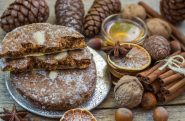 Eine rustikale Präsentation traditioneller Lebkuchen, einige davon gestapelt und teilweise gegessen. Umgeben von Zimtstangen, getrockneten Orangenscheiben, Sternanis, Walnüssen, Haselnüssen, Tannenzapfen und einer kleinen Schale Honig auf einer Holzoberfläche.