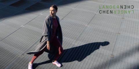 Une personne en tenue de sport et portant un long manteau court sur une surface en caillebotis métallique, projetant une ombre. Dans le coin supérieur droit, on peut voir le texte abstrait vert "LANGERNECHE".