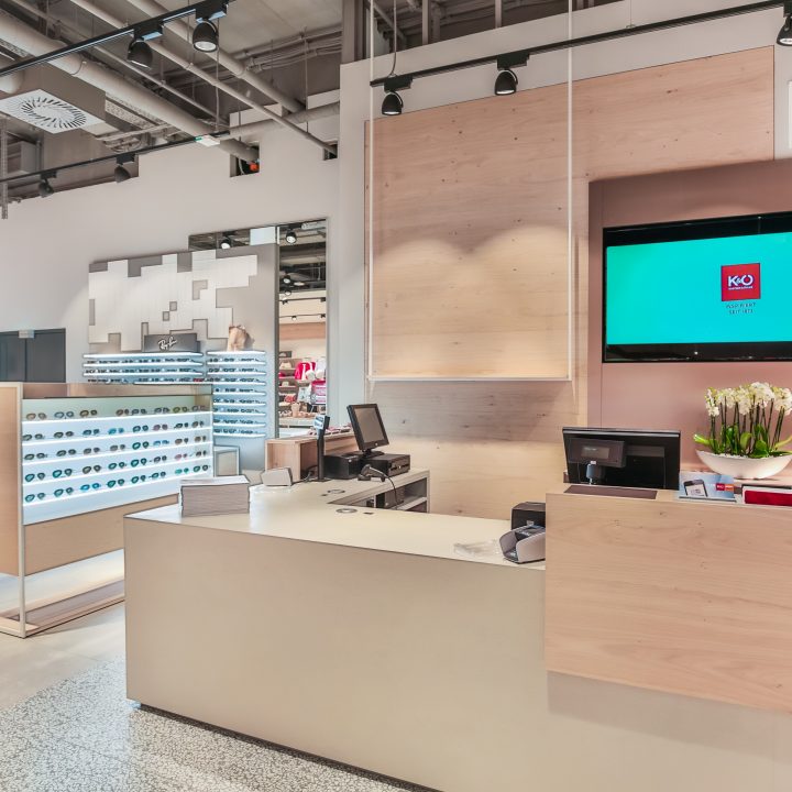 Un intérieur moderne d'un magasin d'optique avec des accents de bois et des présentoirs remplis de montures de lunettes. Sur le côté droit, une caisse enregistreuse avec des écrans d'ordinateur. Au-dessus, un écran de télévision affiche le logo K&O. Un éclairage vif met en valeur le design élégant.
