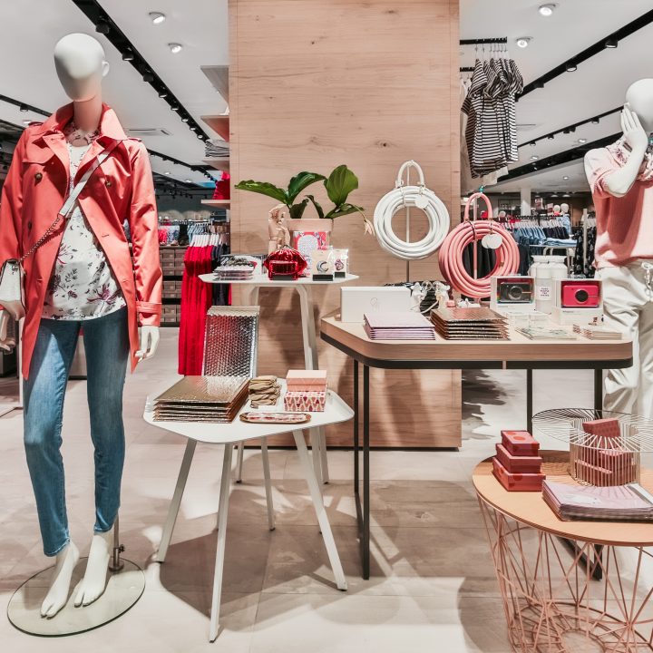 Une vitrine élégante dans un magasin de vêtements avec des mannequins portant des tenues décontractées. Le mannequin de gauche porte une veste rouge, un chemisier imprimé et un jean, tandis que le mannequin de droite porte une chemise rose et un pantalon blanc. Des accessoires sont disposés sur les tables à côté.