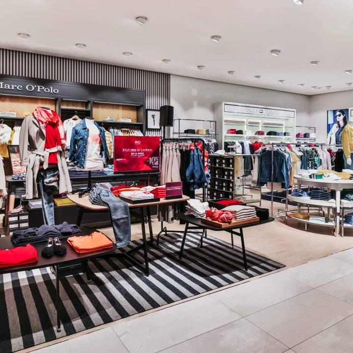 Un intérieur moderne d'un magasin de vêtements avec des rangées de vêtements soigneusement disposées sur des portants et des tables. Une lumière vive met en valeur différents vêtements, notamment des chemises, des vestes et des jeans. Les panneaux de marque pour Mare O’Polo et Tommy Hilfiger sont visibles.