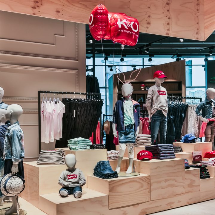 Vitrine d'un magasin de vêtements pour enfants avec des mannequins portant des tenues décontractées, notamment des vestes en jean et des chemises Levi's. Des ballons rouges et blancs flottent au-dessus. Les étagères sont remplies de vêtements pliés et le magasin est bien éclairé.