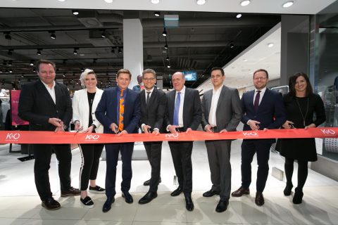 Un groupe de huit personnes se tient dans un magasin et coupe un ruban rouge portant l'inscription "KO" avec des ciseaux. Cela marque l'ouverture d'un nouveau local commercial. Ils portent des vêtements d'affaires et l'ambiance est claire et moderne.