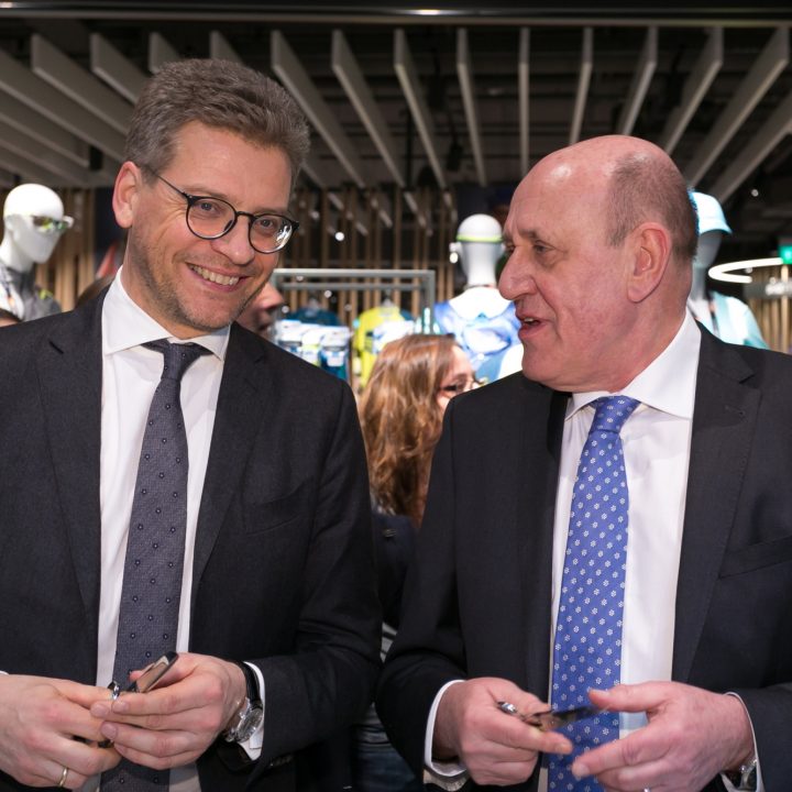 Deux hommes en costume et cravate se tiennent dans un magasin bien éclairé et se sourient. Des mannequins sont visibles en arrière-plan. Ils semblent absorbés par une conversation et tiennent de petits objets dans leurs mains. Derrière eux, on peut voir des gens.