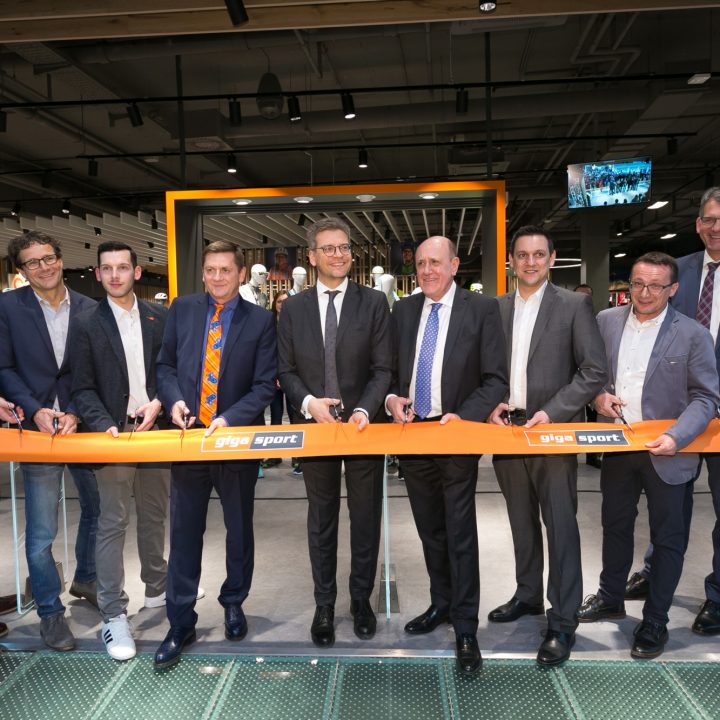 Un groupe de neuf personnes en tenue de ville sourient et tiennent un ruban lors d'une cérémonie d'ouverture de magasin à l'intérieur. Le ruban porte l'inscription "gigasport".