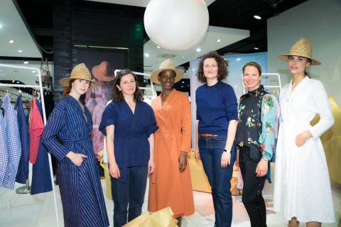 Un groupe de six personnes se tient dans un magasin de détail lumineux. Ils portent des vêtements décontractés et semi-formels. Certains portent des chapeaux de paille. Ils sont entourés de portants à vêtements, et un éclairage et une décoration modernes créent une atmosphère élégante.