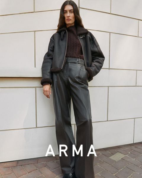 Une personne aux cheveux longs se tient devant un mur carrelé. Elle porte une veste noire et un pantalon foncé taille haute. Sa main gauche est dans sa poche et le mot « ARMA » apparaît en bas de l'image.