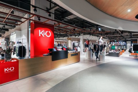 Ein modernes Interieur eines Bekleidungsgeschäfts mit Kleiderständern. Das Geschäft verfügt über eine leuchtend rote Akzentwand mit dem K&Ö-Logo. Es sind Schaufensterpuppen in trendigen Outfits ausgestellt und der Raum hat ein elegantes, offenes Design mit Deckenbeleuchtung.