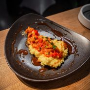 Ein stilvoll angerichtetes Gericht mit einem knusprigen, goldenen Boden, belegt mit einer bunten Mischung aus gewürfeltem Gemüse in einer Tomatensauce. Der Teller ist dunkel und kunstvoll geformt, an den Rändern ist braune Sauce geträufelt.