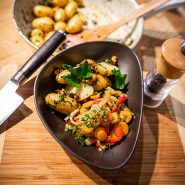 Ein dunkles Gericht mit gerösteten Babykartoffeln, garniert mit Kräutern und bunten Gemüsescheiben wie Paprika. Auf dem Holztisch stehen eine hölzerne Pfeffermühle und ein Messer, im Hintergrund eine Schüssel mit weiteren Kartoffeln.