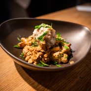 Ein Gericht aus cremigen Champignons, garniert mit frischen Kräutern, wird in einer dunklen, geschwungenen Schüssel serviert. Eine Kugel von etwas, das wie Kartoffelbrei oder eine andere Art von Stärke aussieht, liegt obenauf. Die Schüssel steht auf einem Holztisch.