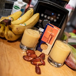 Zwei Gläser Bananen-Smoothie stehen auf einem Tisch, umgeben von frischen Bananen, einer Schachtel Zimtstangen, Datteln und einem Mixer im Hintergrund.