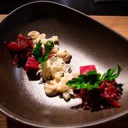 Ein modernes, kunstvolles Gericht auf einem dunklen, asymmetrischen Teller besteht aus kleinen Portionen gewürfeltem rotem Fleisch, zerkleinertem Gemüse, cremigen Klecksen und frischen Kräutern, elegant in minimalistischem Stil auf einer Holzoberfläche angerichtet.