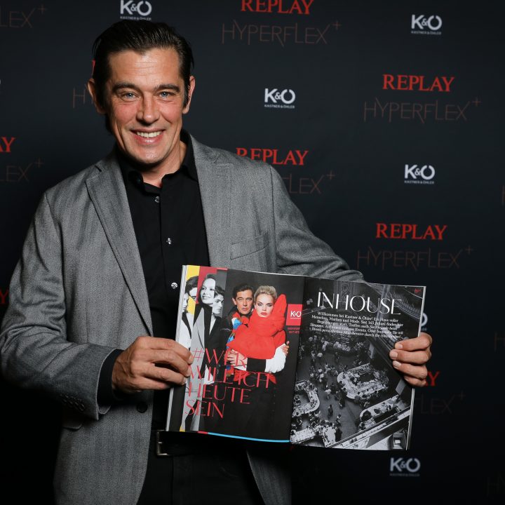 Un homme en costume gris tient à la main un magazine ouvert avec des images de mode et de style de vie. Il se tient devant un fond avec les logos « REPLAY », « HYPERFLEX » et « K-O ». Il sourit à la caméra.