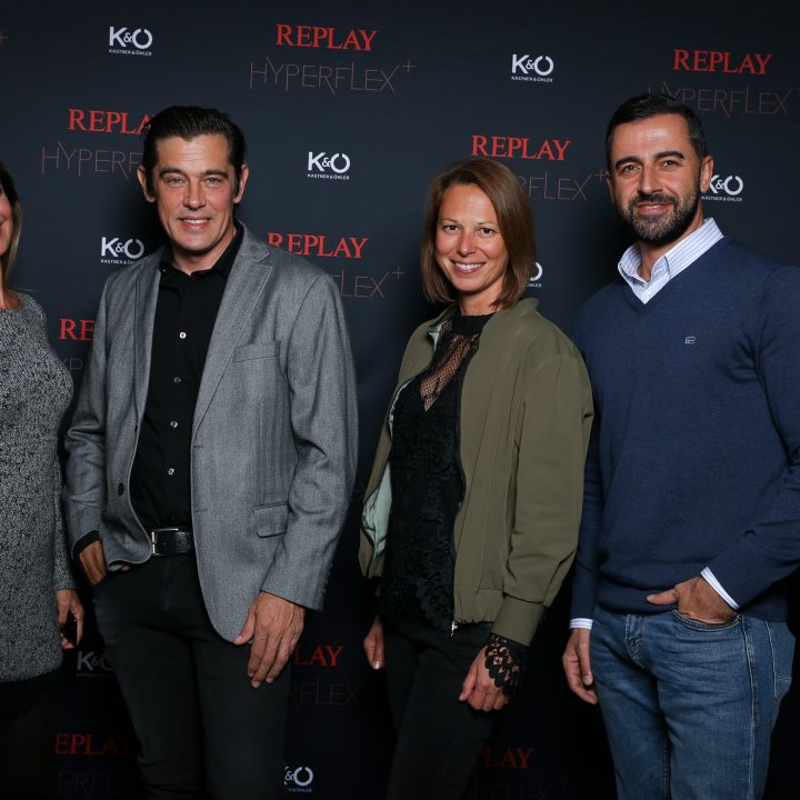 Quatre personnes posent devant une toile de fond avec les logos "Replay Hyperflex" et "K&O". Le groupe est composé de deux femmes, l'une en robe grise, l'autre en veste foncée, et de deux hommes, l'un en costume, l'autre en pull bleu. Ils sourient à la caméra.