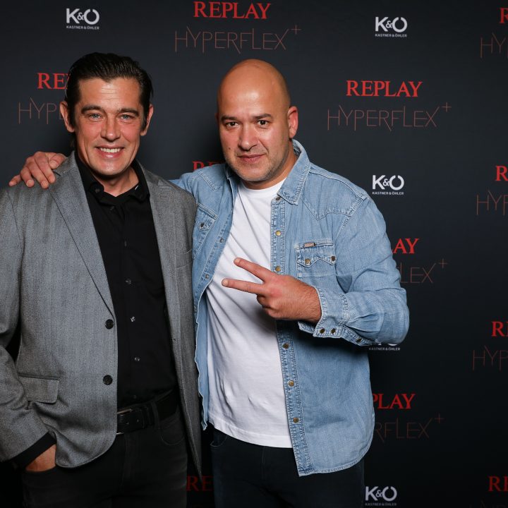 Sur un fond noir avec l'inscription "Replay Hyperflex" et le logo KÜ, deux hommes posent ensemble. L'un porte un blazer gris, l'autre une veste en jean et fait le signe de la paix avec ses doigts.