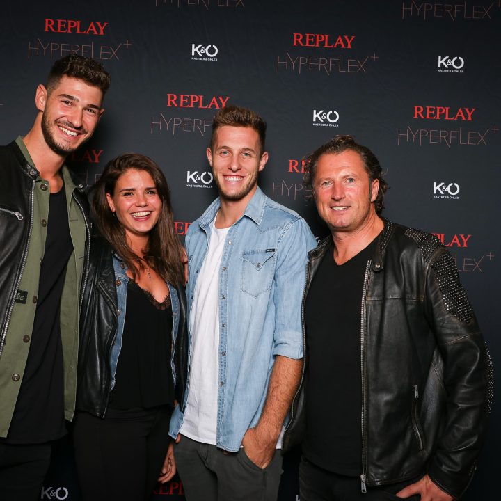 Quatre personnes sourient devant un fond sombre avec les inscriptions « REPLAY HYPERFLEX » et « KÖ ». Elles sont vêtues de manière décontractée, avec des vestes en cuir et en jean.