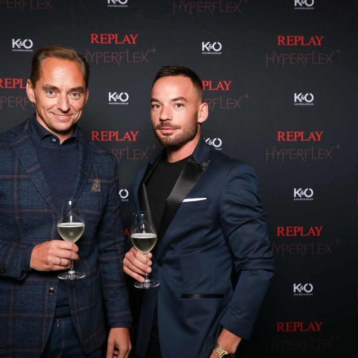 Deux hommes en costume se tiennent debout avec des verres de vin devant un fond noir sur lequel on peut lire des textes rouge et blanc comme "REPLAY HYPERFLEX" et "KO". Ils sourient à la caméra.