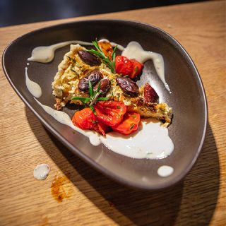 Ein dreieckiges Stück Quiche, garniert mit Kirschtomaten, schwarzen Oliven und Rosmarin, wird auf einem tropfenförmigen schwarzen Teller serviert. Das Gericht wird mit einer weißen Soße beträufelt und auf einem Holztisch platziert.